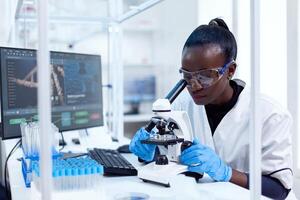 africano médico dentro farmacologia olhando para placa do patoloy dentro paciente amostra usando microscópio. Preto cuidados de saúde cientista dentro bioquímica laboratório vestindo estéril equipamento. foto