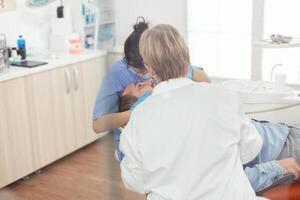 doente paciente sentado em dental cadeira com aberto boca durante estomatologia procedimento. Senior mulher estomatologista e hospital enfermeira com máscaras segurando esterilizado Ferramentas verificação dentes saúde foto