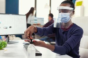 africano mulher Segue segurança precaução durante covid19 usando mão antibacteriano. empresária dentro Novo normal local de trabalho desinfecção enquanto colegas trabalhando dentro fundo. foto