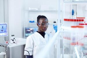 africano cientista segurando teste tubo com azul líquido para médico propósito. Preto investigador dentro estéril laboratório conduzindo farmacologia experimentar vestindo casaco. foto