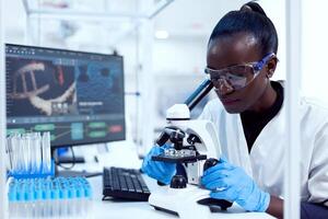 virologista do africano etnia sentado às dela local de trabalho arrumado teste tubos olhando através microscópio. Preto cuidados de saúde cientista dentro bioquímica laboratório vestindo estéril equipamento. foto