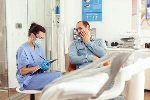 odontologia enfermeira escrevendo notas em prancheta sobre paciente dental problemas esperando para médico ortodontista. assistente sentado dentro estomatológico quarto preparando para dente exame antes cirurgia foto