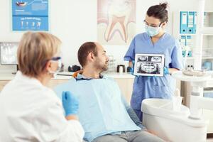 doente homem sentado em dental cadeira olhando às radiografia em digital tábua discutindo com Senior médico estomatologista sobre dor de dente tratamento. médico estomatologia equipe trabalhando dentro odontologia escritório foto