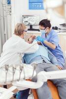 Senior mulher estomatologista analisando com espelho doente paciente dente saúde. médico assistente preparando para limpeza dentes, tratando afetado massa enquanto doente homem sentado em estomatologia cadeira foto