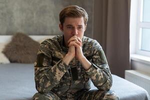 estressado veterano soldado absorta dentro depressivo pensamentos e recordações. triste depressivo jovem militares homem dentro camuflar uniforme sentado em sofá e pensando sobre guerra. ptsd e terapia conceito. foto