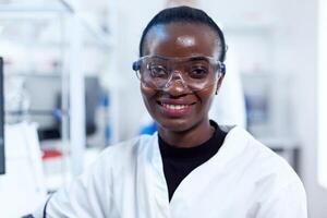 africano saúde químico dentro bioquímica laboratório olhando às Câmera vestindo protetora óculos. multiétnico equipe do pesquisadores trabalhando dentro microbiologia laboratório teste solução para médico propósito. foto