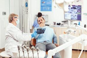 Senior mulher dentista examinando dente radiografia com doente homem discutindo dor de dente durante estomatologia encontro. paciente sentado em dental cadeira dentro hospital odontologia escritório foto