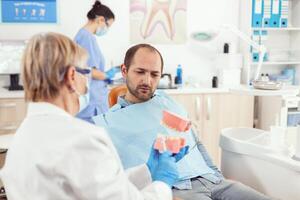 estomatologista Senior mulher segurando médico dente esqueleto explicando dental higiene para homem paciente para evita dente cirurgia enquanto sentado em estomatologia cadeira. médico examinando doente homem foto