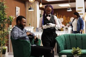 africano americano homem turista sentado dentro hotel lobby pagando para café com Smartphone, fazer sem contato pagamento. amigáveis garçonete segurando pos terminal servindo convidado às luxo recorrer salão área foto