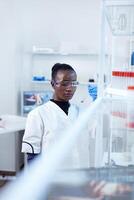 africano cientista examinando azul líquido dentro genético laboratório. Preto investigador dentro estéril laboratório conduzindo farmacologia experimentar vestindo casaco. foto