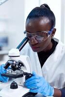 fechar acima do químico do africano etnia fazendo amostra análise olhando através microscópio. Preto cuidados de saúde cientista dentro bioquímica laboratório vestindo estéril equipamento. foto