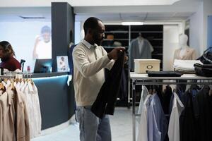 roupas loja cliente explorando prateleira e estante com formal vestem vestuário enquanto compras dentro boutique. homem segurando calças enquanto procurando para Tamanho e escolhendo equipamento dentro sala de exposições foto