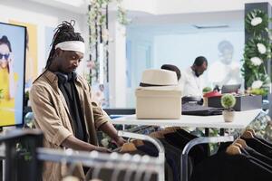 africano americano homem navegando prateleira com suspensão roupas enquanto compras dentro moda boutique. jovem comprador escolhendo à moda equipamento a partir de casual roupa masculina Novo coleção dentro Shopping foto