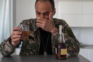 interior tiro do infeliz soldado homem dentro camuflar uniforme e boné guardando mão perto face enquanto sentado em uma sofá, pensamento, tendo pensativo pensativo facial expressão. foto