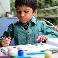 inteligente indiano pequeno Garoto executar polegar pintura com diferente colorida água cor kit durante a verão Férias, fofa indiano criança fazendo colorida polegar pintura desenhando em de madeira mesa foto