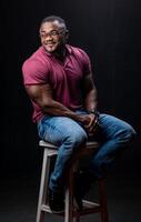 jovem bonito homem dentro vermelho camisa e jeans sentado em cadeira. Atlético muscular masculino posando para Câmera em cadeira. foto