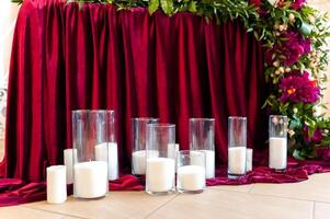 muitos velas dentro vidro estão em pé em a chão em a Borgonha fundo. romântico Projeto para Casamento cerimônia decorado com Borgonha pano e flores com branco velas. foto