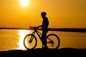 silhueta do uma masculino ciclista com capacete às pôr do sol perto a rio. saudável ativo estilo de vida foto