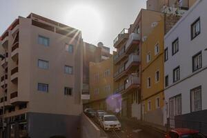 Sol flare sobre urbano rua com moderno apartamento edifícios e estacionado carros, representando cidade vivo dentro los cristãos, tenerife. foto