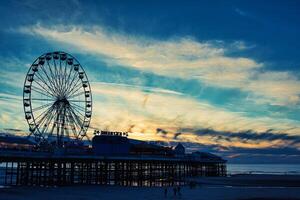 ferris roda em uma cais às pôr do sol com dramático céu e silhuetas do pessoas em a de praia dentro piscina preta, Inglaterra. foto