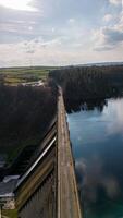 aéreo Visão do uma barragem com uma estrada em principal, dividindo uma floresta e uma calma azul reservatório debaixo uma nublado céu. foto