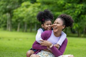 africano americano mãe é jogando pegar carona equitação e abraçando com dela jovem filha enquanto tendo uma verão piquenique dentro público parque para bem estar e felicidade foto