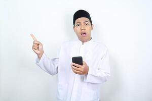 chocado ásia muçulmano homem apontando para cópia de espaço ao lado com segurando Móvel telefone foto
