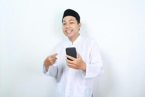 feliz ásia muçulmano homem apontando para Móvel telefone aguarde em mãos com sorridente face isolado foto