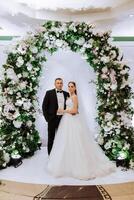 noiva e noivo ficar de pé dentro frente do arco decorado com flores dentro antecipação do Casamento cerimônia foto