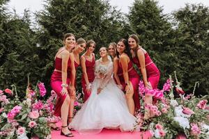 uma morena noiva e dela damas de honra dentro Coincidindo Rosa vestidos estão em pé perto a solene arco. meninas dentro idêntico vestidos estão fazer Fora às uma casamento. Casamento dentro natureza. foto