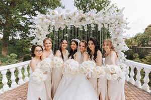 uma morena noiva e dela damas de honra dentro Coincidindo creme vestidos ficar de pé e alegrar com buquês do flores perto a cerimonial arco. Casamento dentro natureza foto