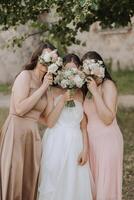 uma morena noiva e dela damas de honra dentro creme vestidos ficar de pé e cobrir seus rostos com buquês do flores meninas dentro Coincidindo vestidos. Casamento dentro natureza foto