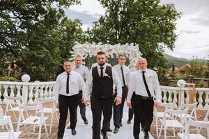 alegre, jovem, enérgico testemunhas do a noivo Próximo para a noivo. amigos felicitar a noivo. a noivo dentro uma colete e dele amigos dentro branco camisas pose perto a Casamento arco. Casamento dentro natureza. foto