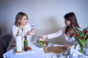 uma Adolescência menina e dela mãe dentro pijamas estão cozinhando e comendo uma fresco verde e tomate salada juntos foto