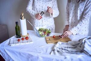 uma Adolescência menina e dela mãe dentro pijamas estão cozinhando e comendo uma fresco verde e tomate salada juntos foto