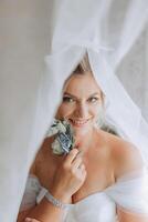 lindo jovem noiva segurando véu dentro branco Casamento vestir, retrato do morena noiva dentro hotel sala, manhã antes casamento. foto