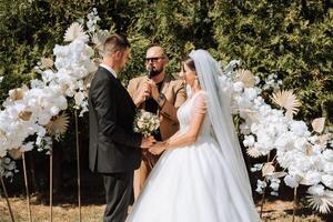 Casamento cerimônia dentro natureza. a noiva e noivo perto a flor arco. mestre do cerimônias dentro Sombrio óculos às uma Casamento durante uma desempenho contra a fundo do a noiva e noivo. foto