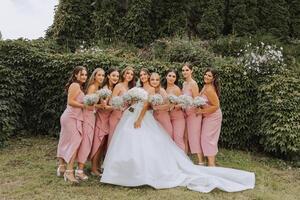 uma morena noiva e dela damas de honra dentro Coincidindo Rosa vestidos estão em pé com buquês do flores dentro seus mãos. meninas dentro idêntico vestidos estão fazer Fora às uma casamento. Casamento dentro natureza. foto