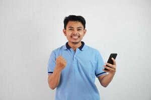 animado ou feliz retrato jovem ásia homem segurando telefone com cerrado punhos vestindo azul pólo t camisa isolado em branco fundo foto