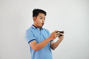 animado ou feliz jovem ásia homem jogando jogos com Smartphone vestindo azul t camisa isolado em branco fundo foto