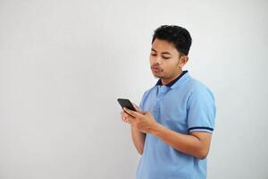 jovem ásia homem em pé segurando uma telefone com a Bravo e desaprovando expressão vestindo azul t camisa isolado em branco fundo foto
