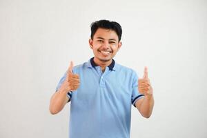 retrato do alegre ásia homem dentro vestindo azul t camisa sorridente e mostrando polegares acima às Câmera isolado sobre branco fundo foto