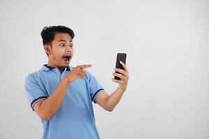 retrato ásia homem chocado segurando telefone e apontando às a telefone com uma dedo vestindo azul pólo t camisa isolado em branco fundo foto