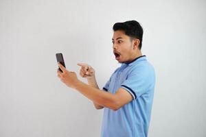 retrato ásia homem chocado segurando telefone e apontando às a telefone com uma dedo vestindo azul pólo t camisa isolado em branco fundo foto