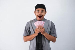 chocado jovem ásia muçulmano homem segurando papel dinheiro presente thr a partir de família Ramadã eid al-fitr Tempo vestindo cinzento muçulmano roupas isolado em branco fundo foto
