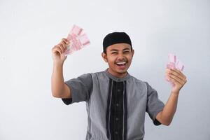 chocado jovem ásia muçulmano homem segurando papel dinheiro presente thr a partir de família Ramadã eid al-fitr Tempo vestindo cinzento muçulmano roupas isolado em branco fundo foto