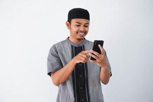 feliz ou sorridente jovem ásia muçulmano homem segurando e apontando em telefone celular vestindo Koko roupas isolado em branco fundo. Ramadã conceito foto