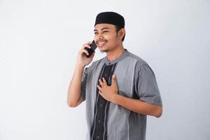 sorridente jovem ásia muçulmano homem segurando peito e segurando telefone chamando de Móvel telefone vestindo Koko roupas isolado em branco fundo foto