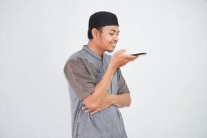 jovem ásia muçulmano homem sorrir falando usando a voz assistente em Móvel telefone vestindo muçulmano roupas isolado em branco fundo foto
