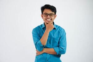 sorridente empregado ásia homem com óculos enquanto segurando a queixo olhando Câmera vestindo azul camisa isolado em branco fundo foto